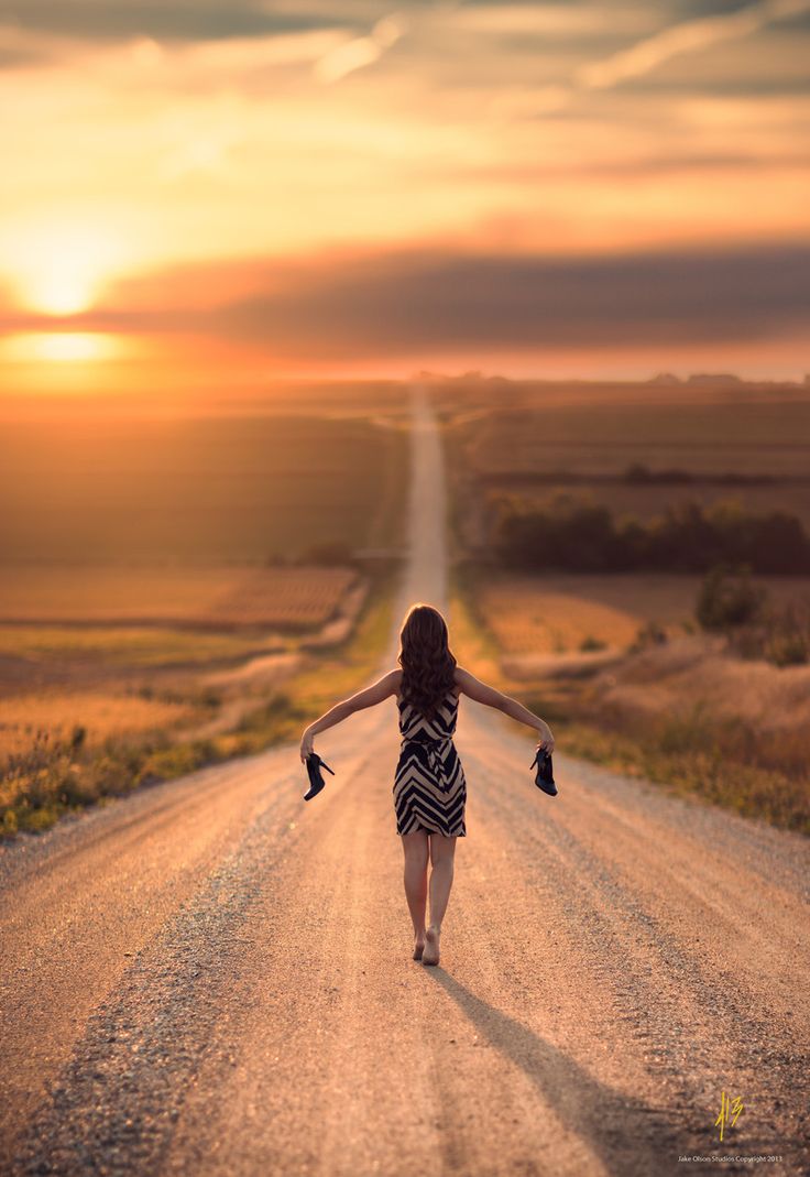 a woman walking down the middle of a dirt road with her arms spread out in the air