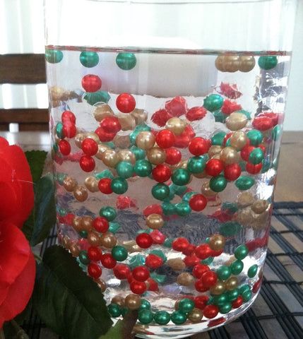 a glass filled with red, green and gold beads next to a flower on a table