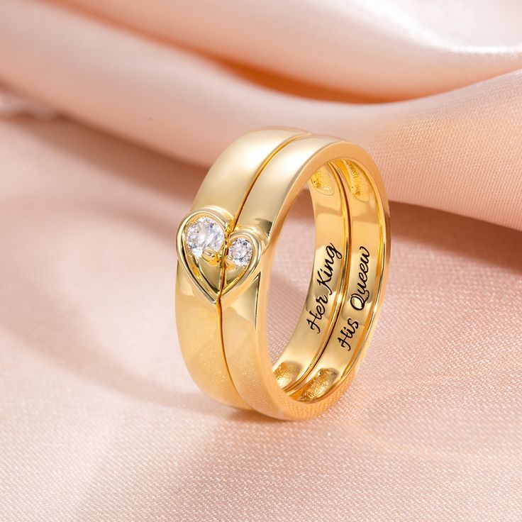 two gold wedding rings with heart shaped diamonds sitting on top of each other, next to a pink cloth