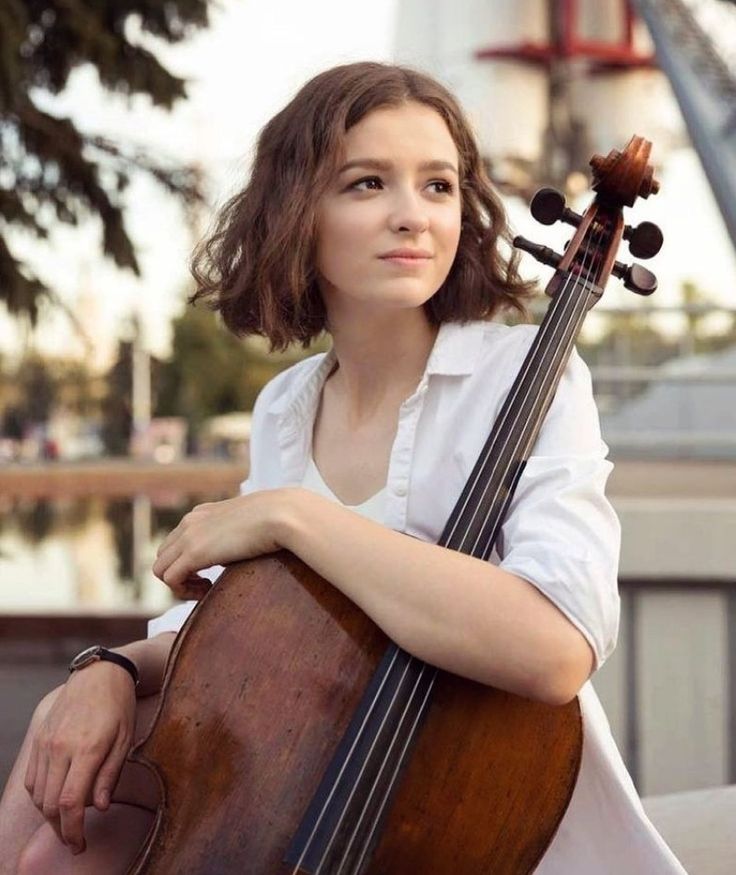 a woman sitting on a bench holding a cello in her hands and looking off into the distance