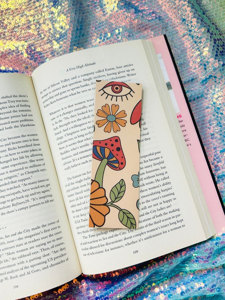 an open bookmark with flowers and eyes on it sitting in front of a colorful blanket