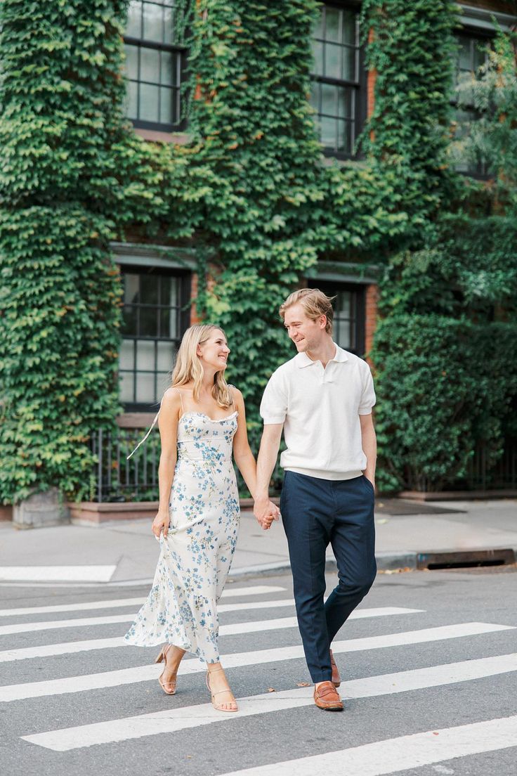 an engaged couple holding hands and walking across the street in front of ivy covered buildings