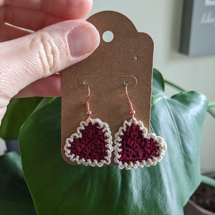 a pair of crocheted heart earrings hanging from a brown card with a green plant in the background