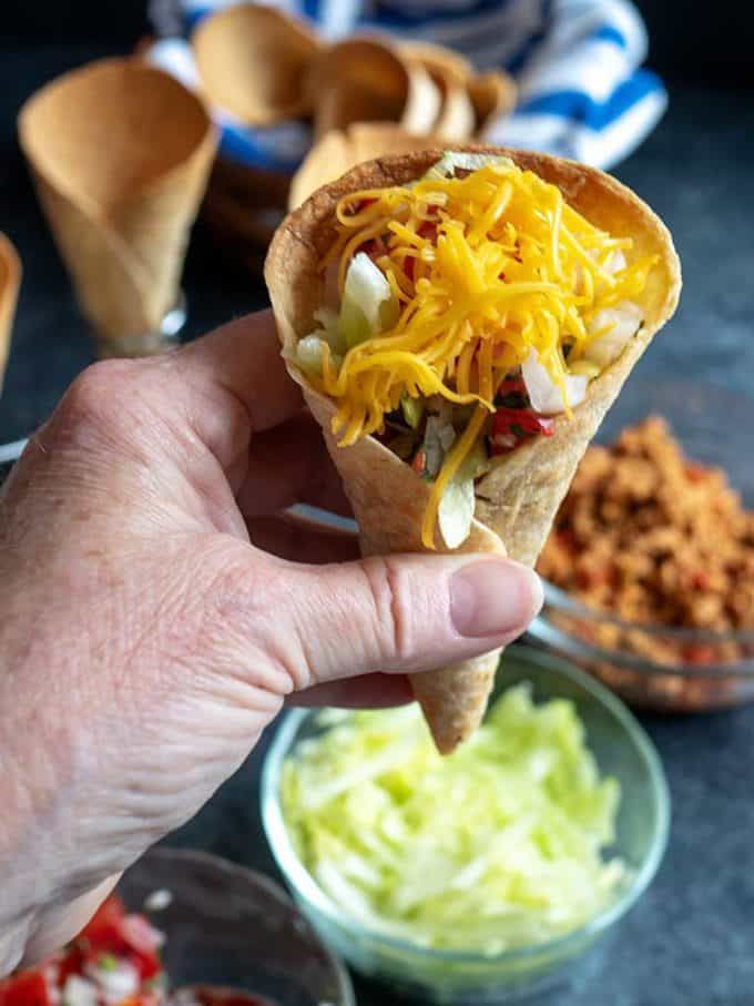 a hand holding up a burrito filled with shredded cheese and toppings next to bowls of other food