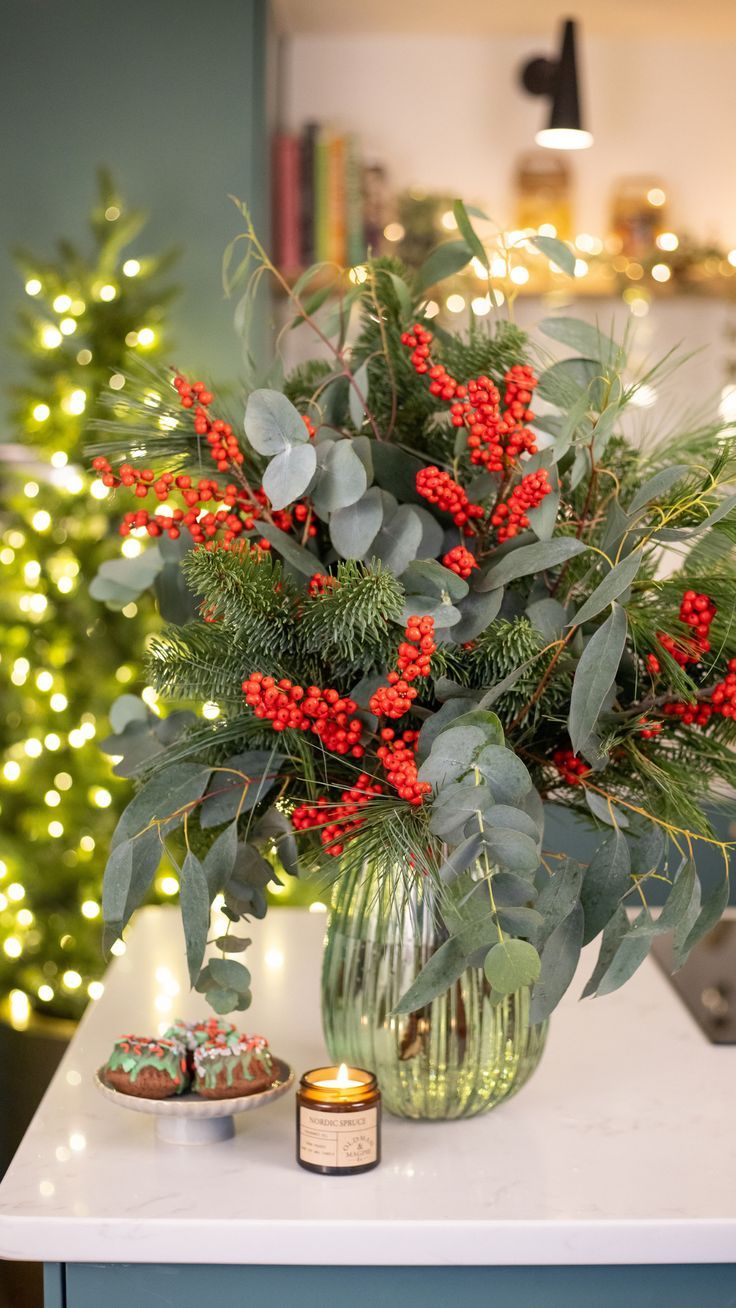 a vase filled with red berries and greenery on top of a table next to a christmas tree