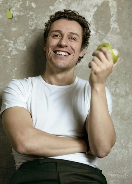 a man holding an apple in his right hand and smiling at the camera while sitting against a wall