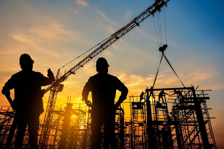 two construction workers silhouetted against the setting sun in front of a building under construction