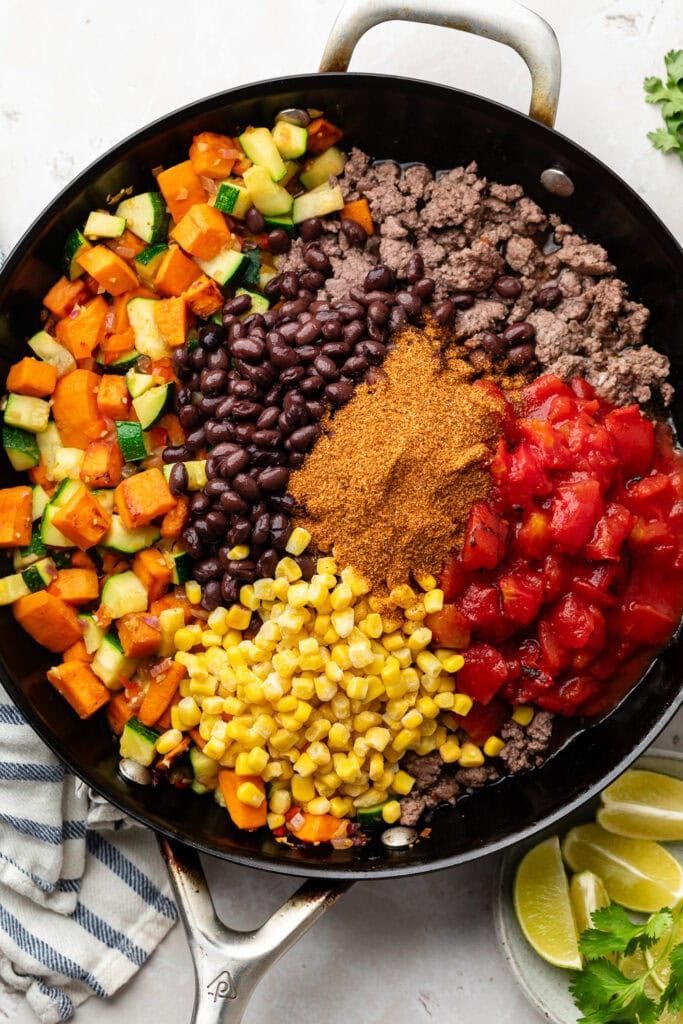 a skillet filled with beans, corn, tomatoes and other veggies next to lime wedges