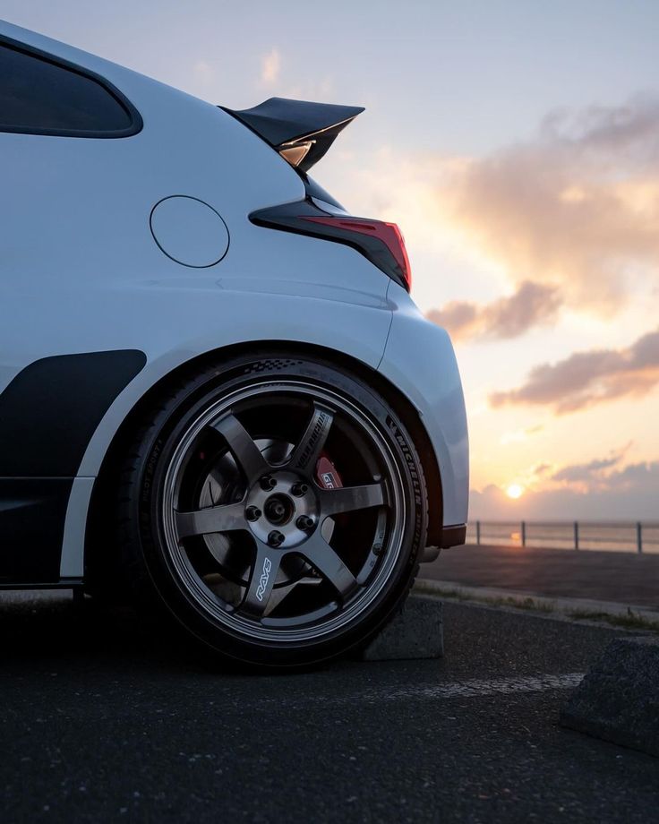 the back end of a white car parked in a parking lot with sunset behind it