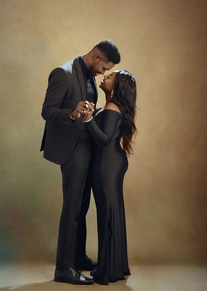 a man and woman standing next to each other in front of a brown background wearing black clothing