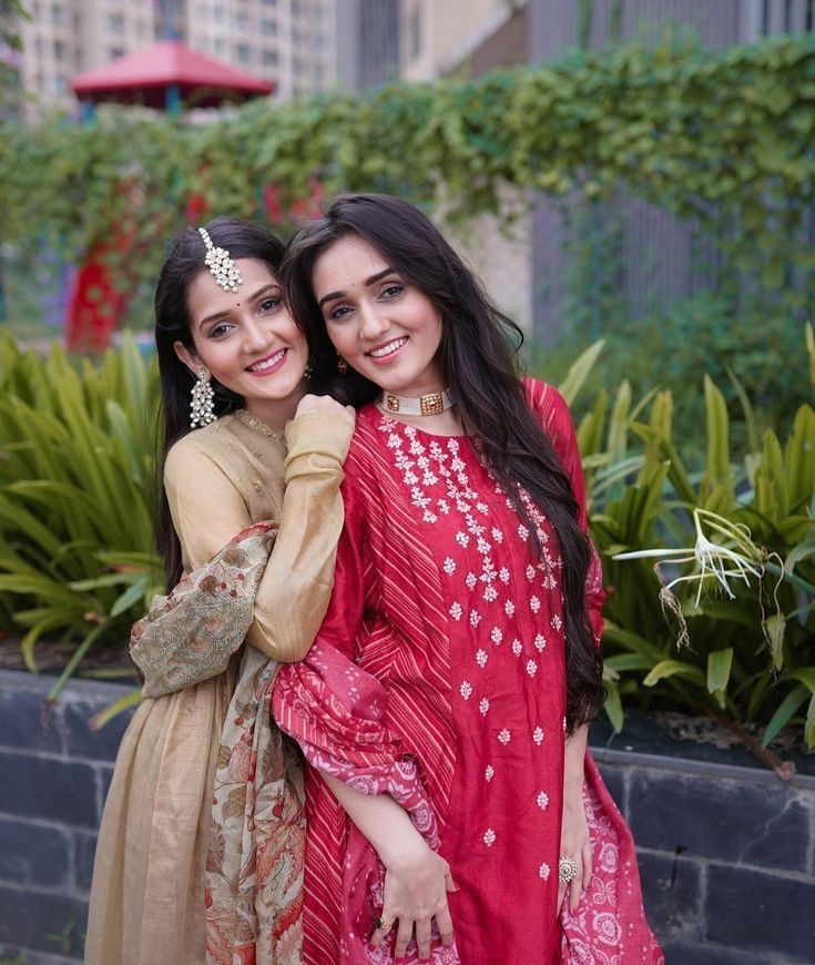 two young women standing next to each other in front of plants and bushes, one holding her arm around the other's shoulder