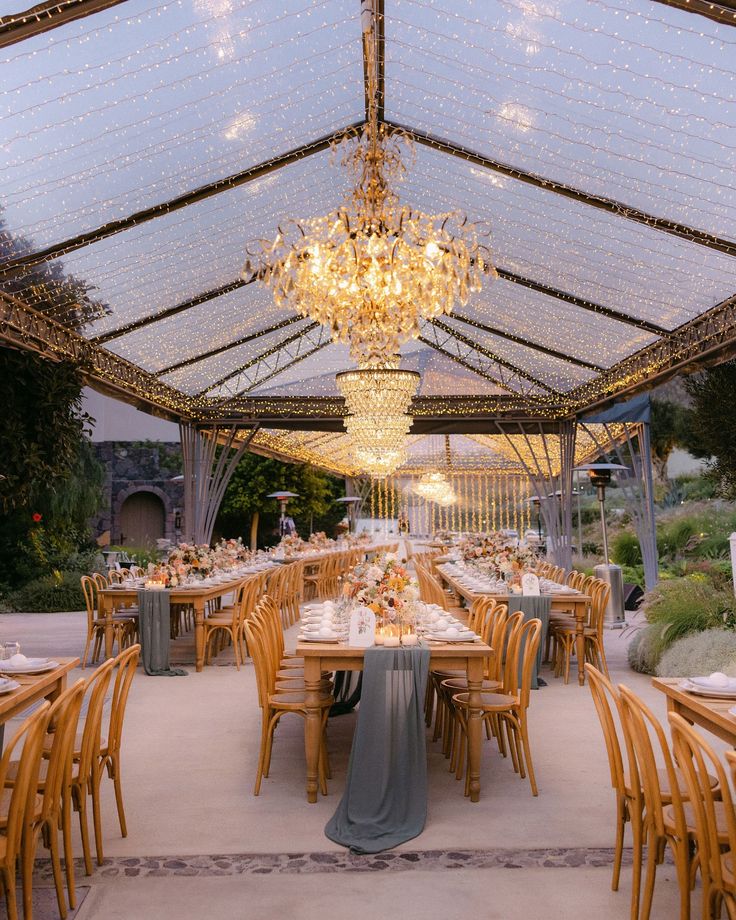 an outdoor dining area with wooden chairs and tables set up for a formal dinner under a glass roof