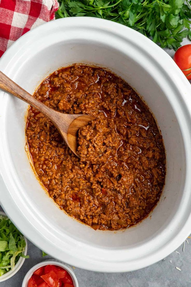 crock pot taco meat in a white bowl with a wooden spoon and tomatoes on the side