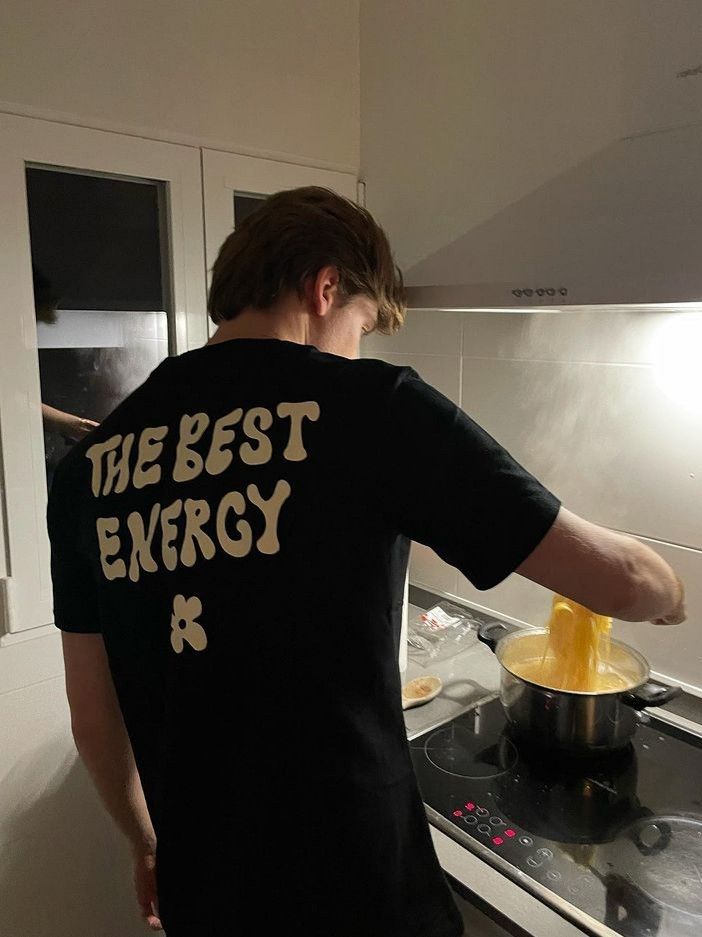 the man is cooking in the kitchen with his hand on the stove top and he is wearing a t - shirt that says the best energy