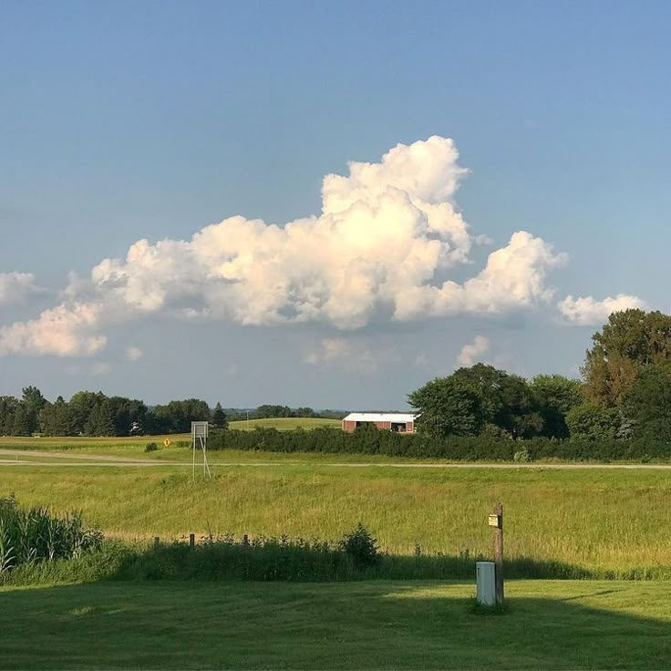 a large field with grass and trees in the background, under a blue sky filled with clouds