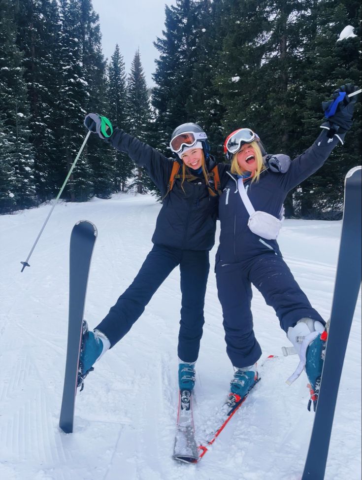 two women standing on skis in the snow with their arms out and one holding her leg up