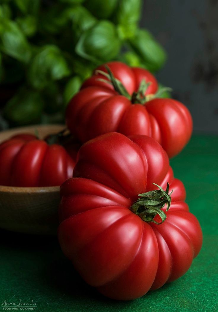 three red tomatoes sitting on top of a green counter next to a wooden bowl filled with basil