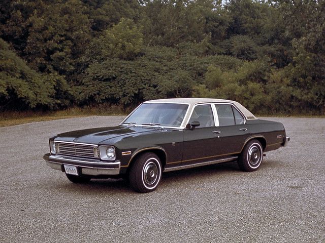 an old black car parked in a parking lot next to some bushes and trees on the other side of the road