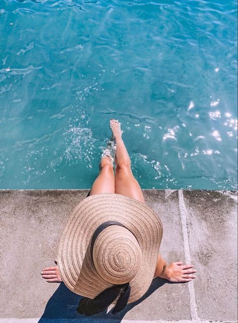 a woman in a hat is sitting by the water with her feet on the ground