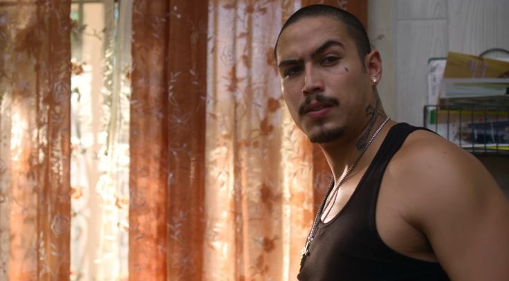 a man in a black tank top standing next to a window with curtains behind him