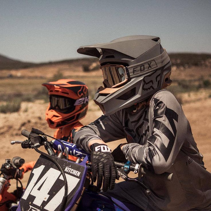 two people on dirt bikes wearing helmets and protective gear, in the middle of an open field