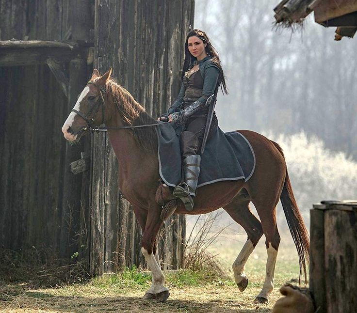 a woman riding on the back of a brown horse next to a wooden structure and dog