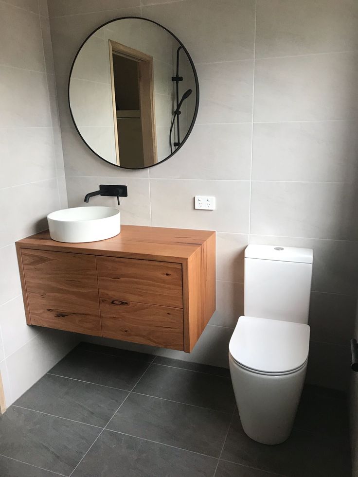 a white toilet sitting next to a wooden sink vanity in a bathroom under a round mirror