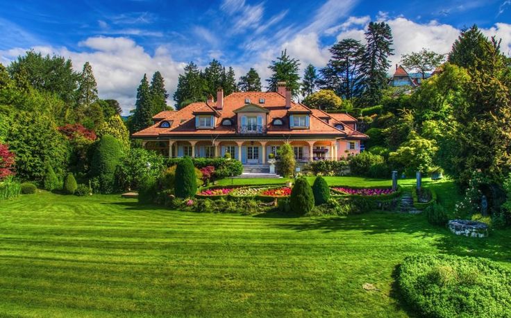 a large house surrounded by lush green trees and flowers in front of a blue sky