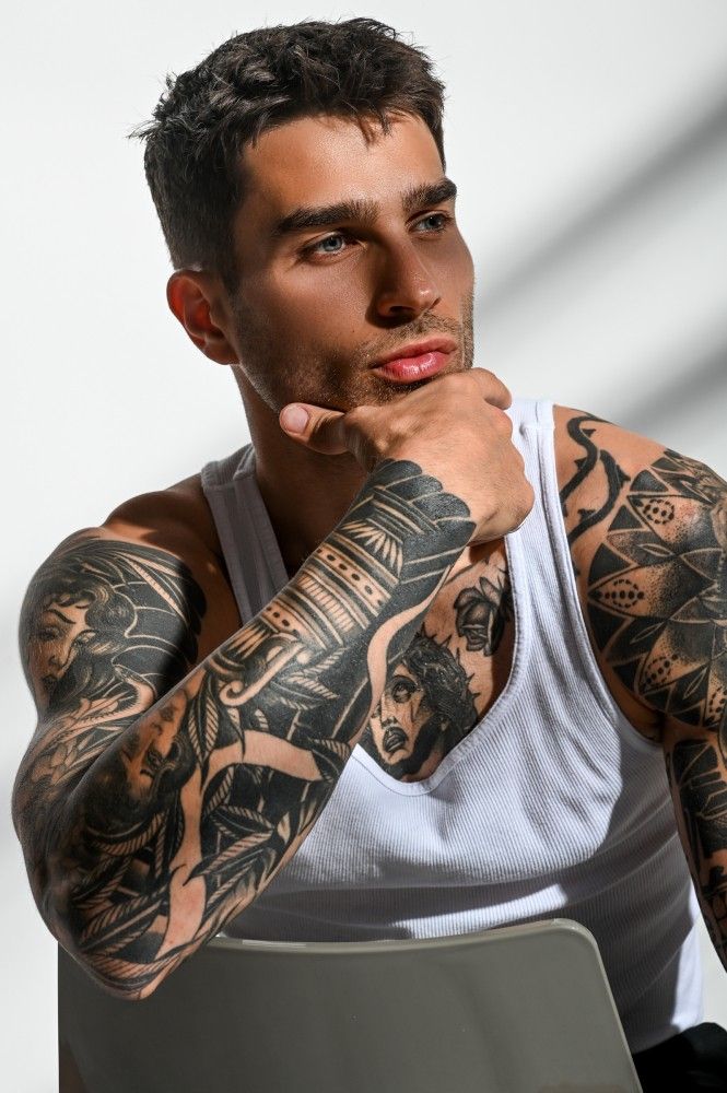 a man with tattoos on his arms and shoulder sitting in front of a white wall