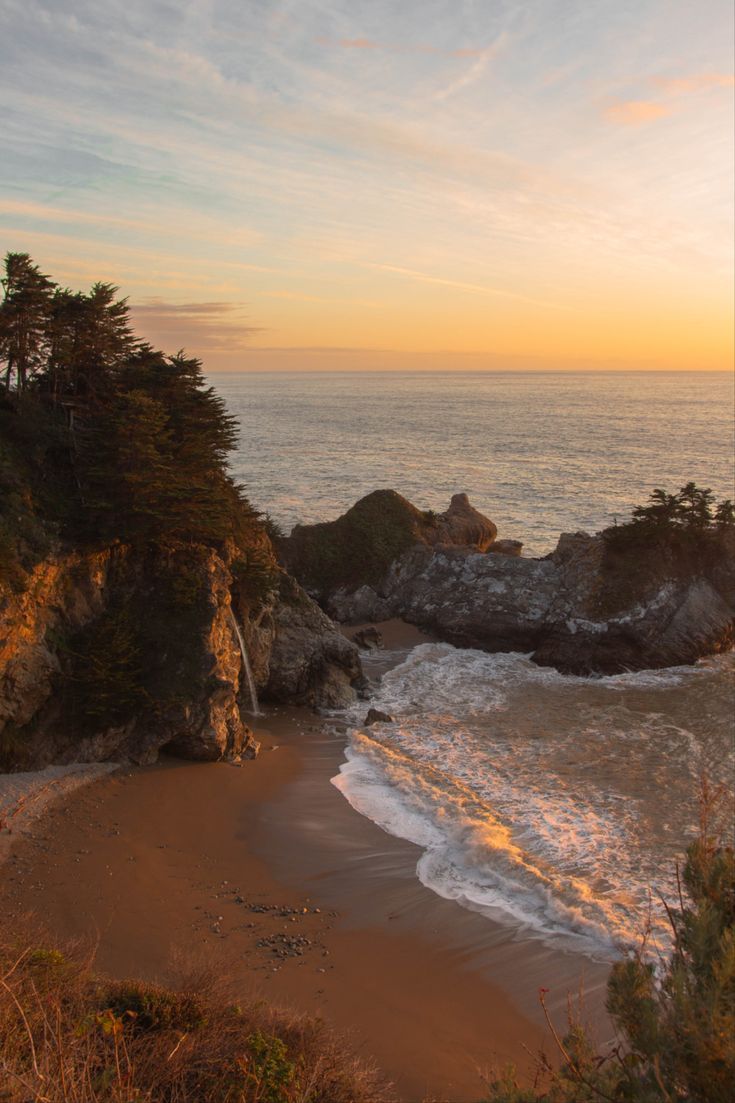 the sun is setting over an ocean with trees on the shore and rocks in the foreground