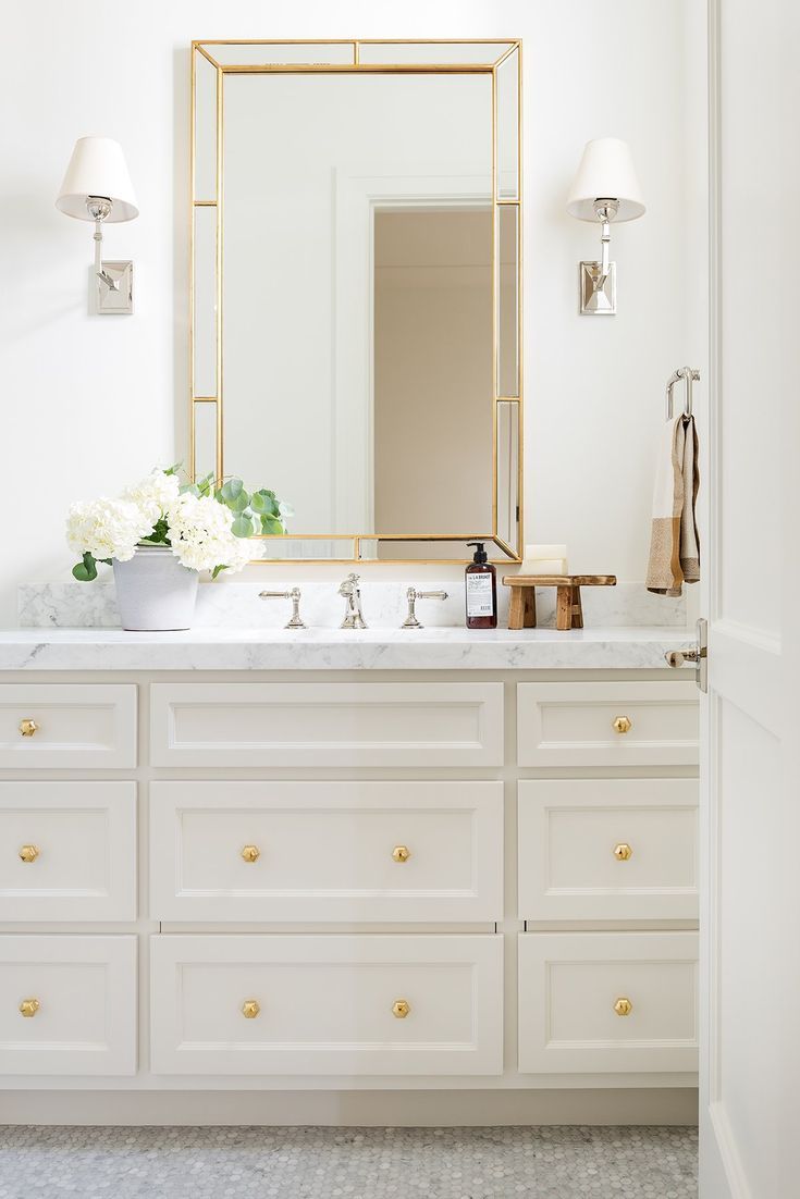 a white bathroom with gold accents and marble counter tops, along with a large mirror above the sink