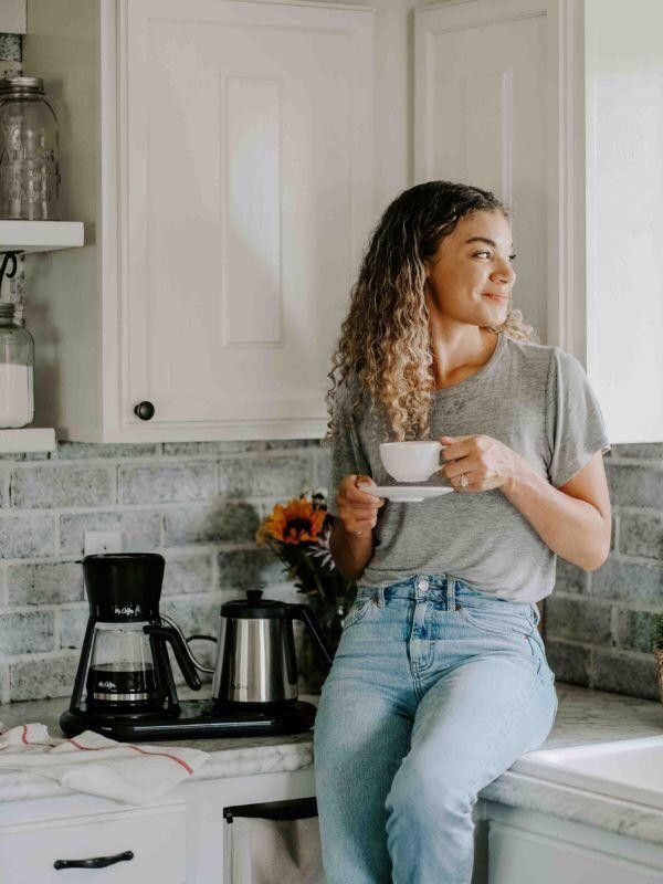 a woman sitting on the kitchen counter drinking from a cup and holding a coffee mug