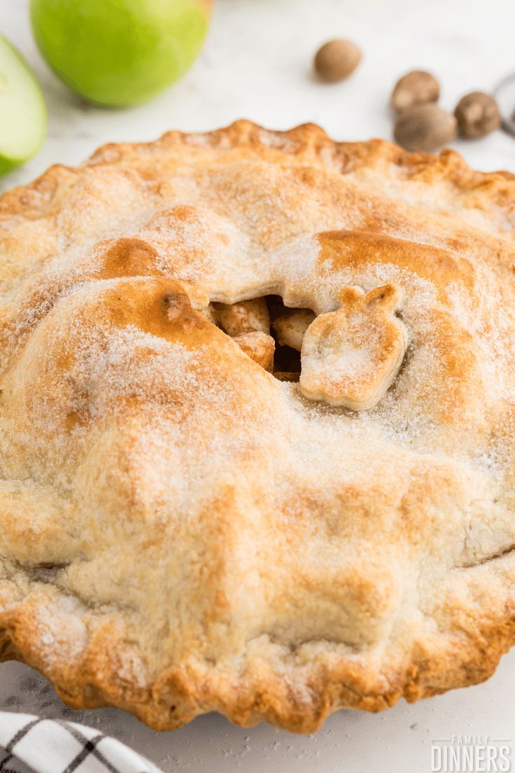 an apple pie with some powdered sugar on top and apples in the back ground