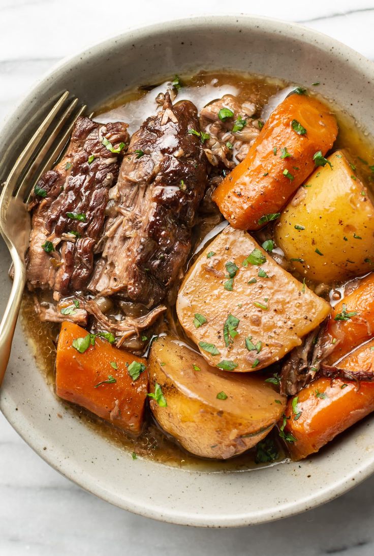 a white bowl filled with roast and potatoes on top of a woven table cloth next to silverware