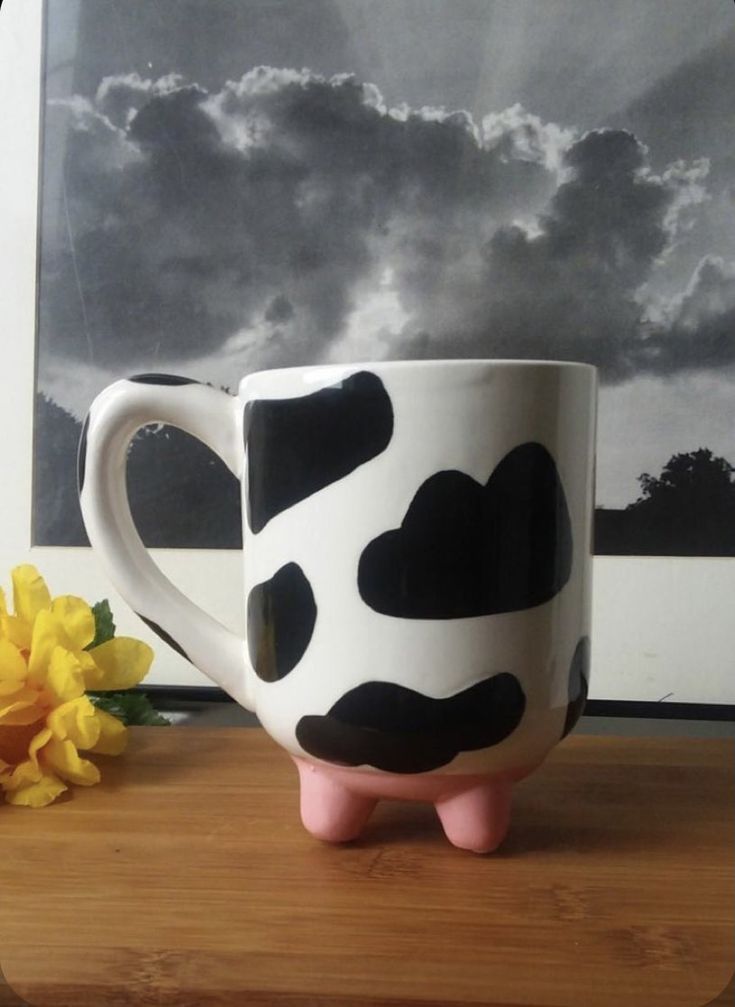 a cow shaped coffee mug sitting on top of a wooden table next to a yellow flower