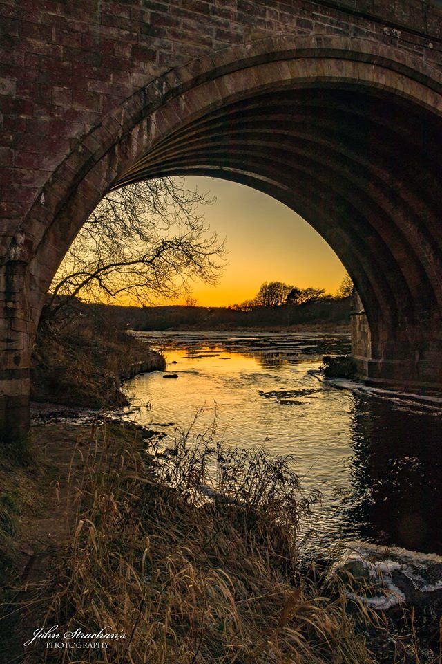 an arched brick bridge over a small river at sunset, with the sun setting in the distance