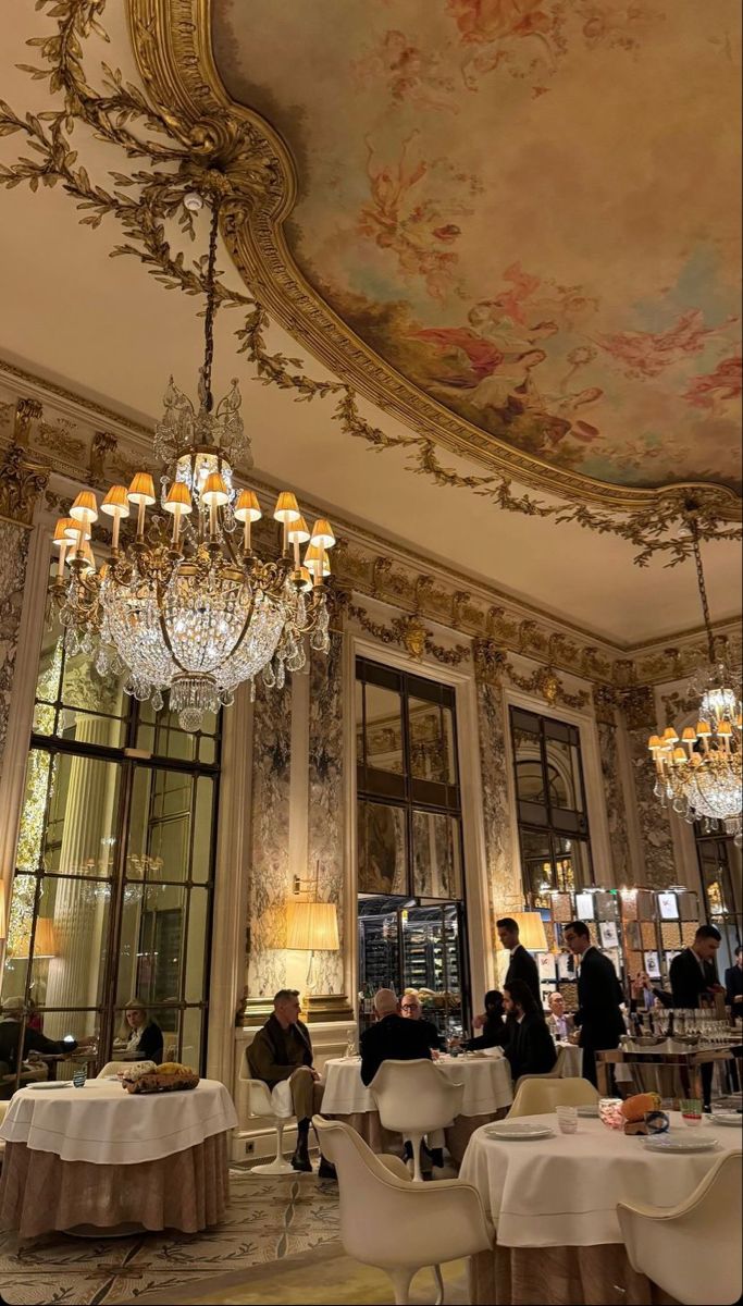 people are sitting at tables in a fancy restaurant with chandeliers and paintings on the walls