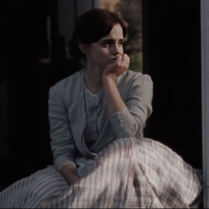 a woman sitting on top of a bed next to a window with her hand under her chin