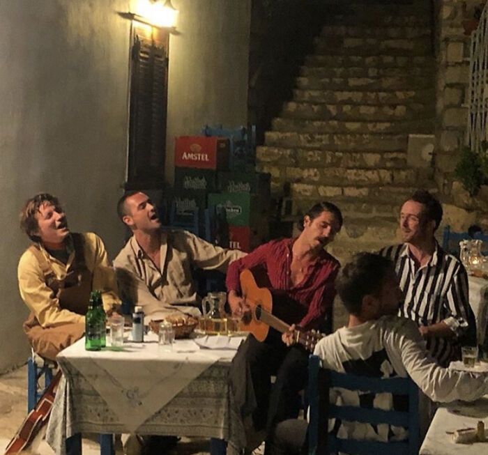 five men sitting around a table with drinks and food on it while one man plays the guitar