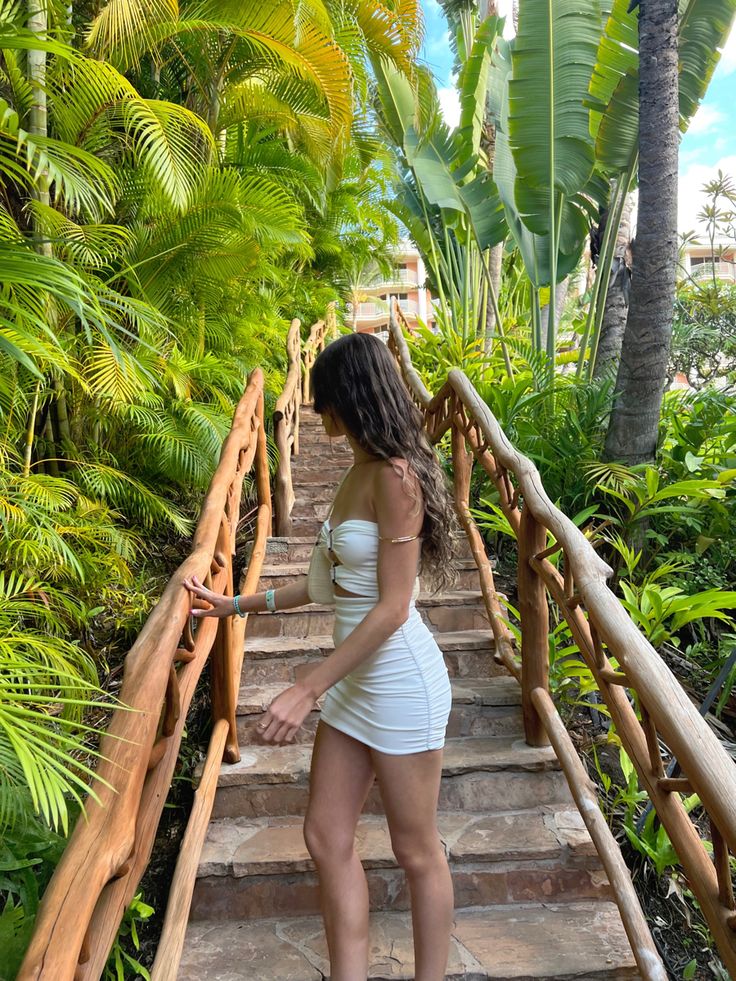 a woman in white dress standing on stairs with palm trees behind her and looking down at the ground