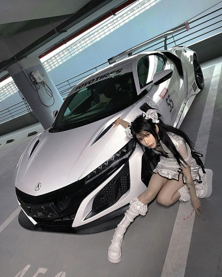 a woman leaning on the hood of a silver sports car in a parking lot next to a white and black car