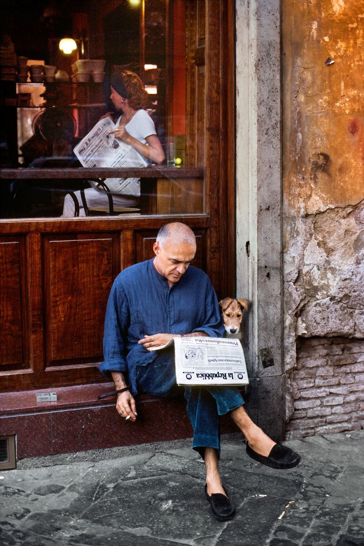 a man sitting on the sidewalk reading a newspaper with his dog in front of him