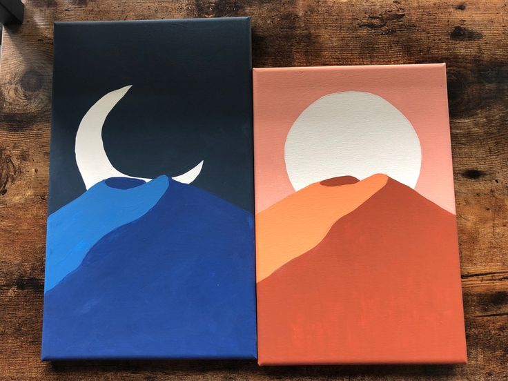 two canvases with mountains and moon on them sitting on a wooden table next to each other