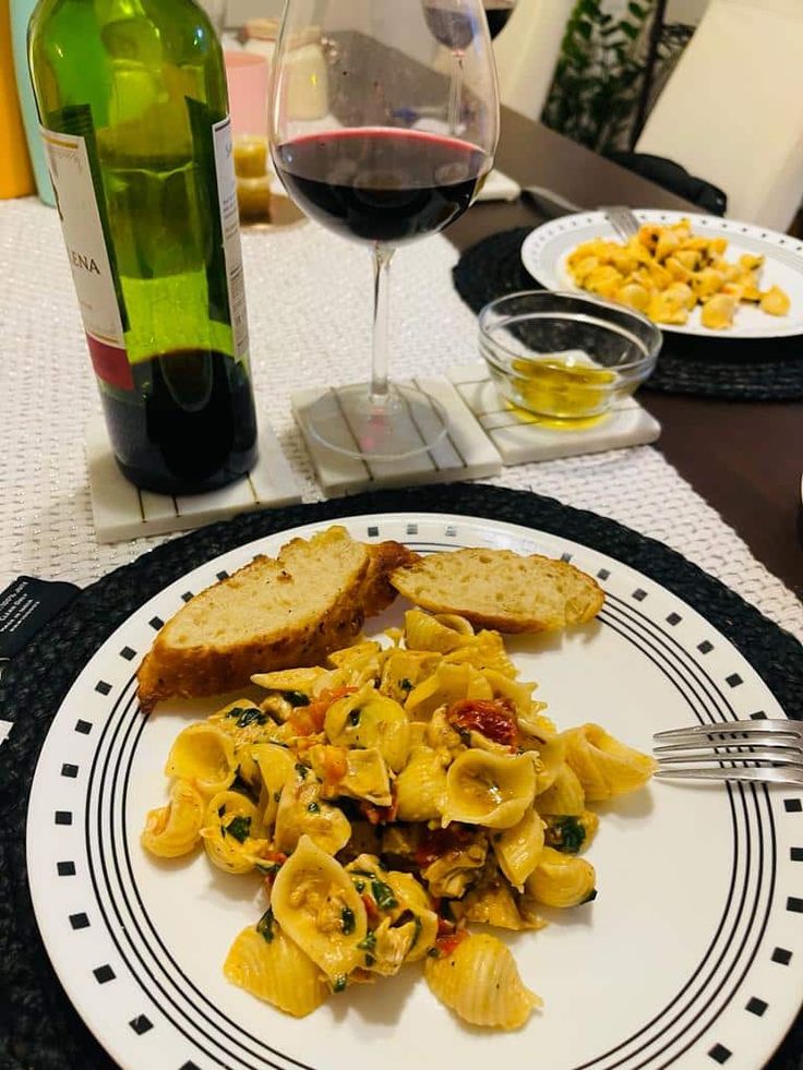 a plate with pasta and bread on it next to a glass of red wine,