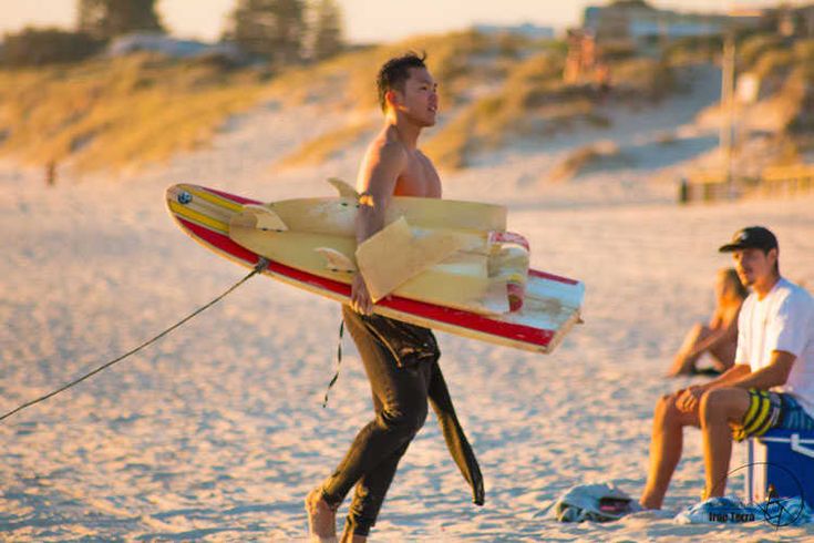 a man holding a surfboard while walking on the beach with other people sitting around