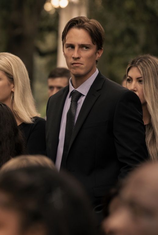 a man wearing a suit and tie standing in front of a group of people with long blonde hair