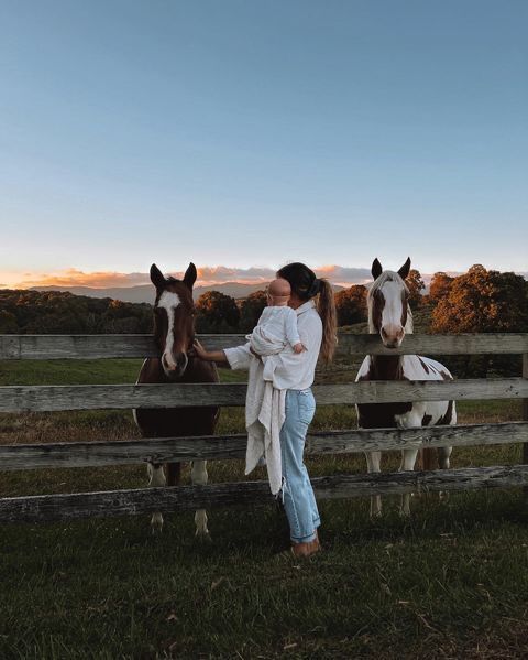 a woman standing next to two horses in a fenced in area with grass and trees