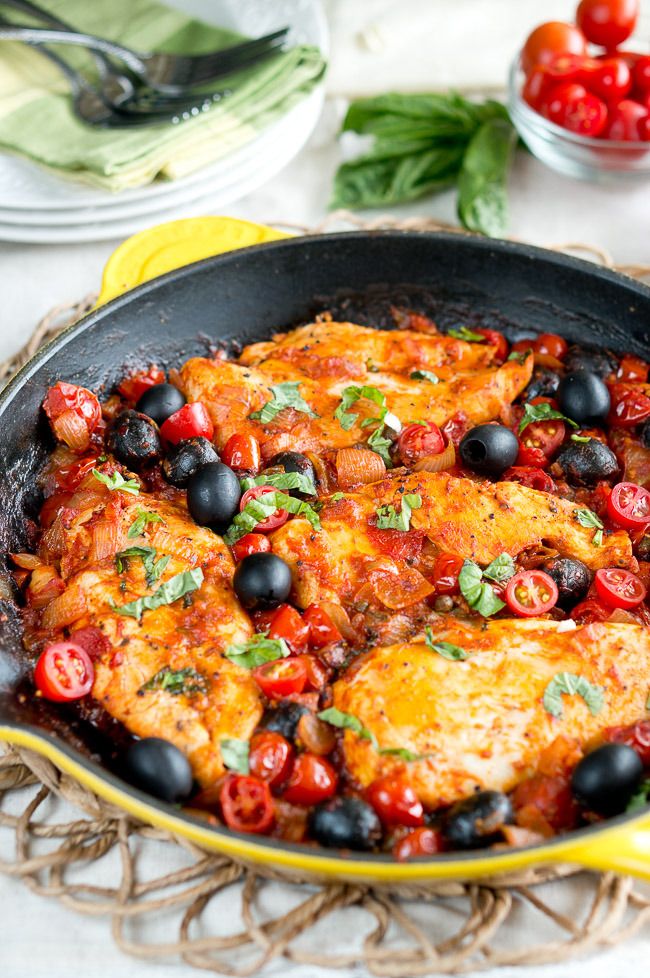 a pan filled with chicken and olives on top of a table next to tomatoes