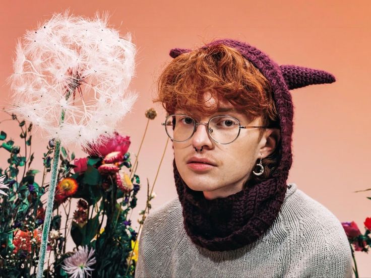 a man with red hair and glasses wearing a knitted cat hat next to flowers