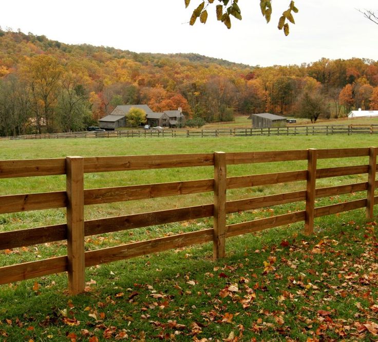 a wooden fence in front of a green field and trees with fall leaves on the ground